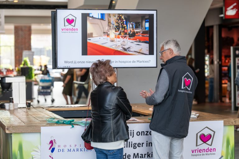 Een vrijwilliger van stichting de vrienden van hospice de markies geeft uitleg aan een voorbijganger tijdens een wervingsactie voor meer vrijwilligers.